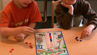 Foto von 2 Kindern, die das NIM-Spiel spielen. Auf dem Tisch liegt das Spielbrett und bereits einige Plättchen. Das Kind links hat rote Plättchen, das Kind rechts blaue.
