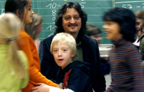 Foto einer Lehrperson vor einer Tafel, umgeben von lachenden Schülerinnen und Schülern.